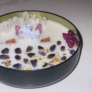 Op de afbeelding: Een zwarte kaars pot met een witte was bovenkant. De kaars is versierd met heldere kwarts kristallen, amethist stenen, gedroogde rozenblaadjes en gouden vlokken.