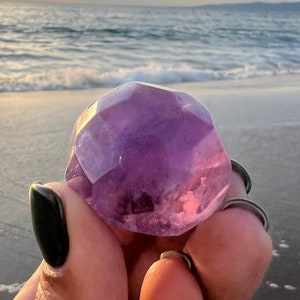 May include: A large, faceted amethyst crystal with a purple hue, held in a hand against a background of a beach and ocean.