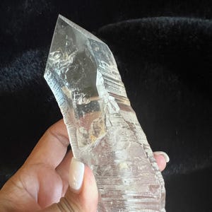 Puede incluir: Un cristal de cuarzo transparente con una punta y una superficie rugosa y texturizada. El cristal se sostiene en una mano sobre un fondo oscuro. El cristal es una gema natural y en bruto.