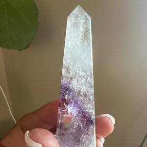 Può includere: Una punta di cristallo di ametista trasparente con sfumature viola e iridescenti. Il cristallo ha la forma di una piramide e una superficie liscia e lucida.