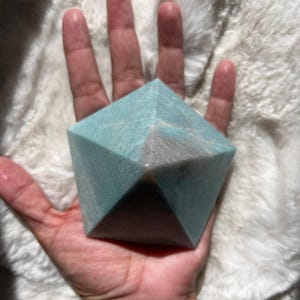 以下が含まれることがあります： 幾何学的な、多面的なターコイズ色のクリスタル。クリスタルは手で持たれており、背景は柔らかく白いふわふわした素材です。クリスタルの形状は五角柱で、淡い青緑色をしています。