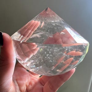 Op de afbeelding: Een heldere, facetgeslepen kristal in een diamantvorm. Het kristal wordt in een hand gehouden en de facetten vangen het licht op, waardoor een sprankelend effect ontstaat. Het kristal is een decoratief item, mogelijk voor weergave of meditatie.