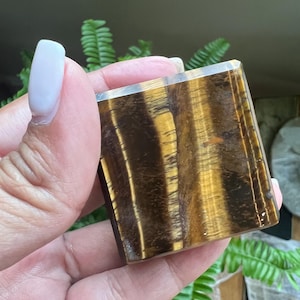 May include: A square-shaped tiger's eye gemstone, showcasing a rich blend of golden-brown and dark brown hues. The polished surface reveals a chatoyant effect, with visible bands and patterns. The gemstone is held in a hand, with a blurred green background.
