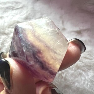 Puede incluir: Un cubo de cristal de fluorita translúcido y multicolor. El cubo muestra bandas de tonos morados, amarillos y claros. El cristal se sostiene en una mano, mostrando sus superficies pulidas y su belleza natural. Este espécimen mineral es ideal para coleccionistas.