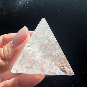 May include: A clear, triangular crystal pyramid. The pyramid is held in a hand and appears to be made of quartz or a similar translucent material. The edges are sharp, and the crystal has internal fractures.