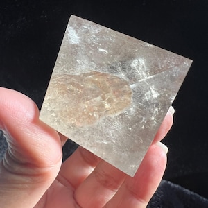 Puede incluir: Un cristal transparente de forma cuadrada con una inclusión marrón claro en el centro. El cristal se sostiene en una mano sobre un fondo oscuro. El cristal es translúcido y parece ser un objeto decorativo.