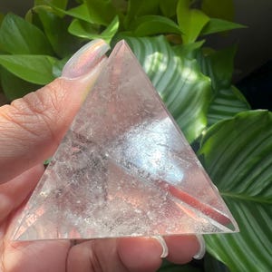May include: A clear, triangular-shaped quartz crystal pyramid with visible internal fractures and inclusions. The stone has a smooth, polished surface and a pointed apex, reflecting light through its transparent, crystalline structure.
