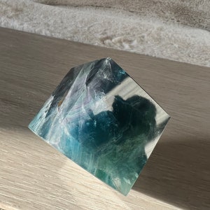 May include: A green and blue fluorite pyramid crystal on a light wood surface.