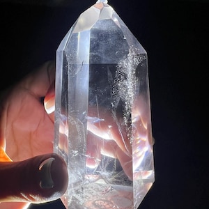 Könnte beinhalten: Ein klarer Quarz-Kristallpunkt mit einer facettierten, geometrischen Form. Der Kristall wird vor einem dunklen Hintergrund gehalten, wobei sich das Licht auf seinen Oberflächen spiegelt. Der Kristall ist etwa 15 cm hoch.