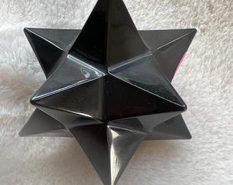 May include: A black, geometric star-shaped object with sharp, pointed edges. The star has a glossy finish and is set against a white, textured background. The object's design suggests a decorative or artistic purpose.