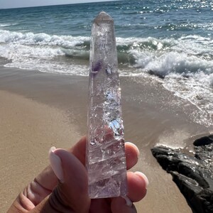 May include: A clear quartz crystal point with purple amethyst inclusions. The crystal is held in a hand against a background of a sandy beach and ocean waves.