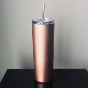 Puede incluir: Un vaso de acero inoxidable color oro rosa con tapa transparente y una pajita plateada. El vaso est&aacute; sobre una superficie negra.