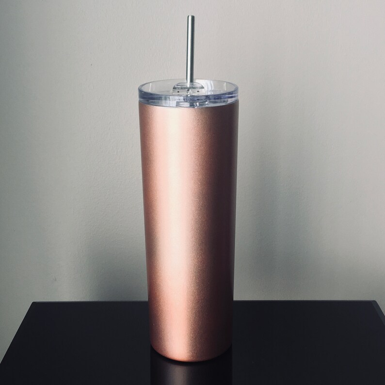 Puede incluir: Un vaso de acero inoxidable color oro rosa con tapa transparente y una pajita plateada. El vaso est&aacute; sobre una superficie negra.