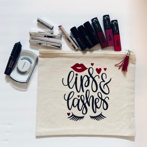 Könnte beinhalten: Eine weiße Kosmetiktasche aus Canvas mit einem roten Reißverschluss und einem roten Quastenanhänger. Die Tasche ist mit den Worten "lips & lashes" in Schwarz bedruckt, dazu ein roter Lippenstift und Wimpern. Die Tasche ist gefüllt mit verschiedenen Kosmetikartikeln, darunter Lippenstifte, Mascara und Wimpernverlängerungen.