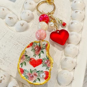 May include: A handmade keychain featuring a painted oyster shell with a floral and heart design. The keychain includes a red heart charm, pink and white beads, and a gold-tone clasp. The shell is edged in gold.