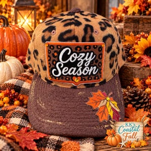 May include: A baseball cap with a tan and black leopard print crown and a solid brown brim. A rectangular patch on the front features the text Cozy Season in white script, accented with orange rhinestones. A fabric maple leaf applique is on the brim.