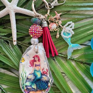May include: A mermaid-themed keychain featuring a painted oyster shell with a mermaid illustration, a red tassel, and beaded accents. Additional charms include a starfish, seashell, and a mermaid tail. The keychain is attached to a silver clasp.