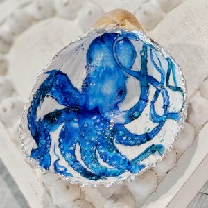 May include: A seashell dish featuring a blue watercolor octopus design. The octopus is painted on the inside of the shell, which has a silver-painted rim. The shell is set on a white, heart-shaped tray with small, white decorative elements.