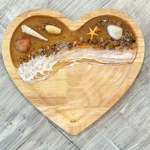 May include: Heart-shaped wooden tray with a beach scene. The tray features a sandy resin base with seashells, a starfish, and a wave design. The tray is made of light-colored wood.