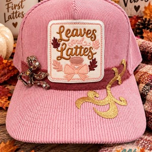 May include: A pink corduroy baseball cap featuring a central rectangular patch with the embroidered text Leaves and Lattes, surrounded by leaf motifs and a bow. The cap is adorned with a jeweled bow on the left and gold embroidery on the right.