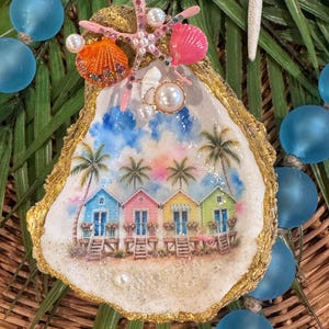 May include: A decorative oyster shell featuring a painted beach scene with four colorful houses, palm trees, and a blue sky. The shell is embellished with a pink starfish, small orange and pink seashells, and white pearls, with gold-painted edges.
