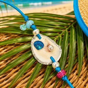May include: A blue cord bracelet featuring a central oyster shell adorned with a small blue seashell charm, a white pearl, and a blue seashell decal. The cord is accented with white, light blue, and pink beads, finished with a silver-toned clasp.