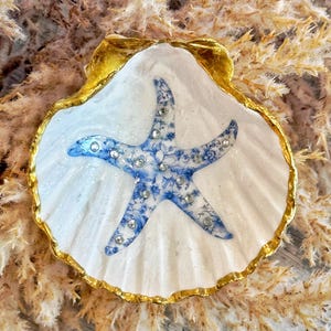 May include: A decorative seashell trinket dish. The white shell has a gold-painted rim and a blue and white floral starfish design in the center. The starfish is embellished with small, clear rhinestones. The dish is a coastal-themed decorative item.