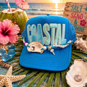 May include: A bright blue trucker hat featuring the word COASTAL in large, light blue embroidered letters. The hat is decorated with a string of white pearls, a small oyster shell, and a silver-toned cross charm attached to the front brim.