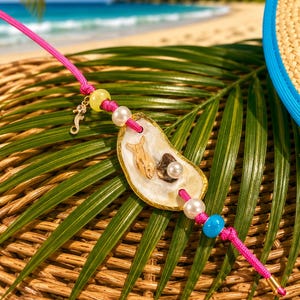 May include: A bright pink cord necklace featuring a central oyster shell pendant adorned with a small fish charm, a dark stone, and white pearls. The cord is accented with additional yellow, white, and blue beads and a gold-toned seahorse charm.