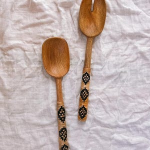 May include: Two wooden salad servers. One is a spoon, the other a fork. Both have light brown wooden heads and handles wrapped with a black and tan woven pattern. The servers are on a white, textured surface.