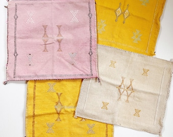 May include: Four square decorative pillows in shades of pink, yellow, and cream. Each pillow features a geometric embroidered design in contrasting colors. The pillows have tassels on the corners and are arranged on a white surface.