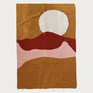 May include: A rectangular woven wall hanging featuring a stylized landscape. The design includes a large white circle representing the sun, a dark red mountain range, a pinkish-beige foreground, and a golden-brown background.