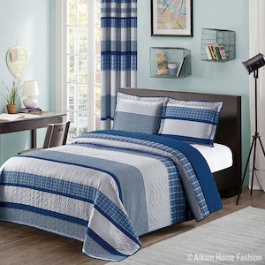 May include: A blue and gray striped quilt with a plaid pattern. The quilt is on a bed with two pillows. The bed is in a bedroom with a window, a lamp, and a desk.