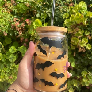 May include: A glass tumbler with a bamboo lid, filled with a light brown beverage and ice. The glass is decorated with black bat and yellow star decals. A silver straw is inserted into the drink.
