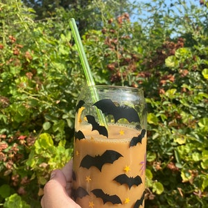 May include: A clear glass with a coffee-colored beverage, decorated with black bat and yellow star decals. A green straw is inserted into the drink. The glass is held in front of a green leafy background.