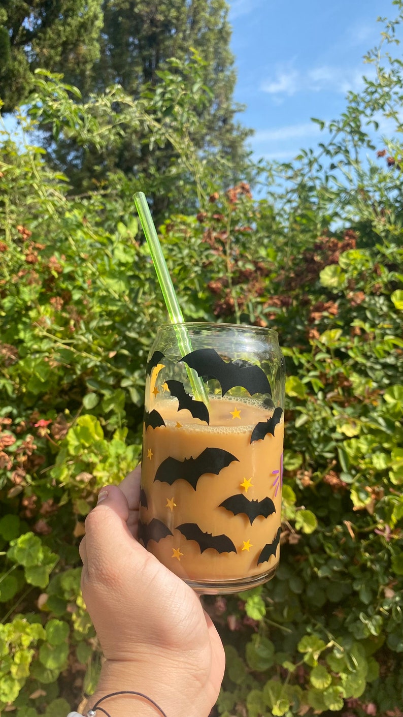 May include: A clear glass with a coffee-colored beverage, decorated with black bat and yellow star decals. A green straw is inserted into the drink. The glass is held in front of a green leafy background.