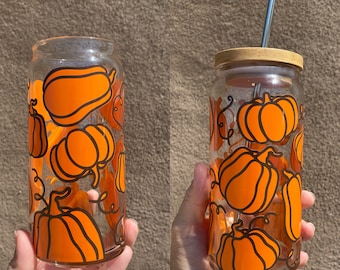 May include: Clear glass tumbler decorated with orange pumpkin designs and black outlines. The tumbler features a bamboo lid and a silver straw. The pumpkins vary in shape and size, with vines. Ideal for autumn.