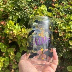 May include: A clear glass can-shaped tumbler decorated with black bats, yellow stars, and a crescent moon. The word "EEK!" is written in purple. The glass is held in front of a green leafy background.