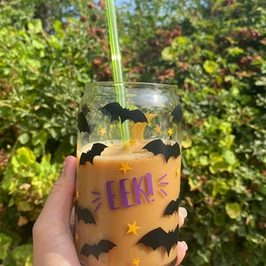 May include: A clear glass can-shaped cup filled with a light brown beverage, decorated with black bat and yellow star decals. The cup has the word "EEK!" in purple. A green straw is inserted into the drink.