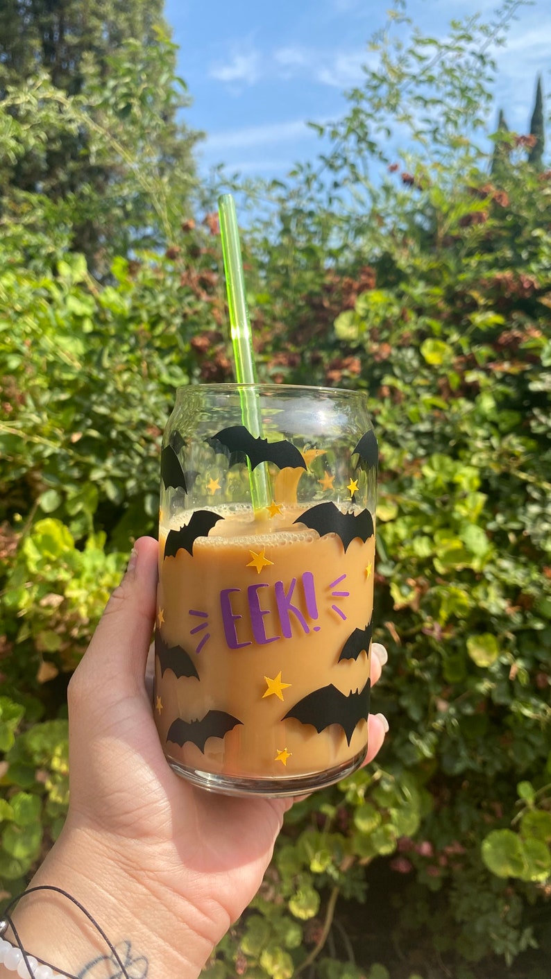 May include: A clear glass can-shaped cup filled with a light brown beverage, decorated with black bat and yellow star decals. The cup has the word "EEK!" in purple. A green straw is inserted into the drink.