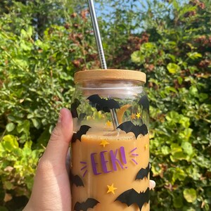 May include: A glass jar with a bamboo lid and a stainless steel straw, filled with a light brown beverage. The jar is decorated with black bat and yellow star stickers, and the word "EEK!" in purple. The jar is held in a hand.
