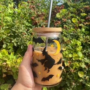 May include: A clear glass beverage cup with a bamboo lid and a stainless steel straw. The cup is decorated with black bats, stars, a yellow crescent moon, and a witch on a broomstick. The cup contains a light brown beverage.