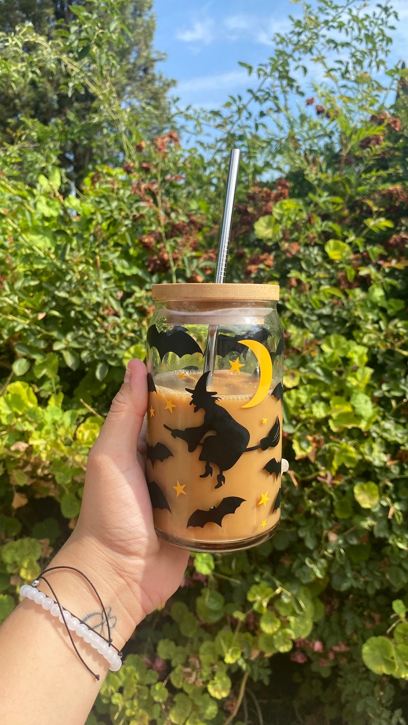 May include: A clear glass beverage cup with a bamboo lid and a stainless steel straw. The cup is decorated with black bats, stars, a yellow crescent moon, and a witch on a broomstick. The cup contains a light brown beverage.