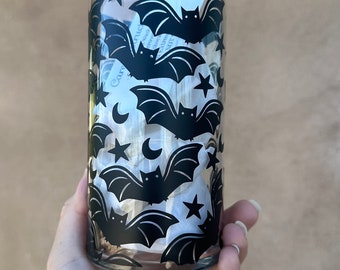 May include: A clear glass can-shaped tumbler decorated with black bat silhouettes, stars, and crescent moons. The Halloween-themed design is repeated around the glass. The tumbler is held in a hand.
