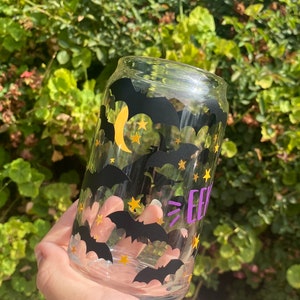 May include: A clear glass can-shaped drinking glass decorated with black bats, gold stars, and a yellow crescent moon. The word "EEK" is printed in purple. The glass is held in front of a green leafy background.