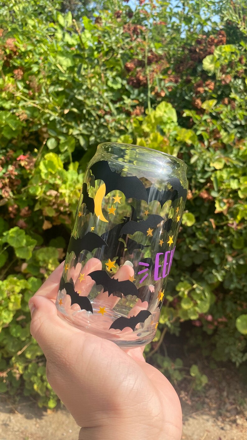 May include: A clear glass can-shaped drinking glass decorated with black bats, gold stars, and a yellow crescent moon. The word "EEK" is printed in purple. The glass is held in front of a green leafy background.