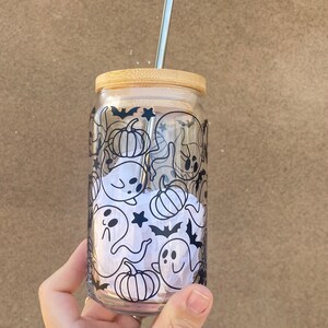 以下が含まれることがあります： 竹製の蓋と銀色の金属ストローが付いた透明なガラスタンブラー。ガラスには、幽霊、カボチャ、コウモリ、星のハロウィーンをテーマにした白黒模様が施されています。タンブラーは手で持たれています。