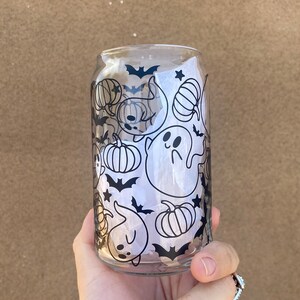 以下が含まれることがあります： ハロウィーンをテーマにしたデザインの、透明なガラス製の缶型カップ。デザインは、カボチャ、コウモリ、星、幽霊の黒いイラストが特徴です。カップは手で持たれています。