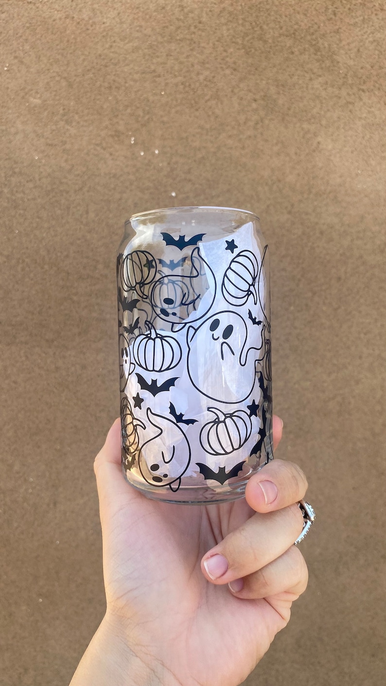 以下が含まれることがあります： ハロウィーンをテーマにしたデザインの、透明なガラス製の缶型カップ。デザインは、カボチャ、コウモリ、星、幽霊の黒いイラストが特徴です。カップは手で持たれています。