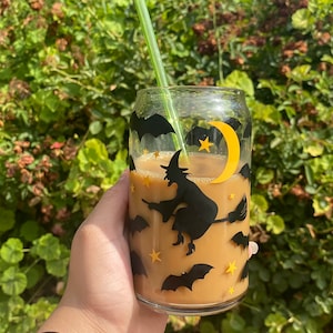 May include: A clear glass cup with a Halloween theme, filled with a light brown beverage and a green straw. The cup is decorated with black bats, stars, a witch on a broom, and a yellow crescent moon.